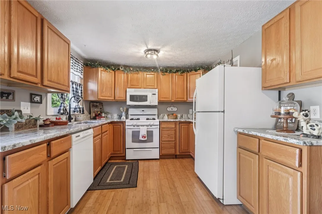 Kitchen with white appliances, light wood finished floors, a textured ceiling, brown cabinets, and light stone counters