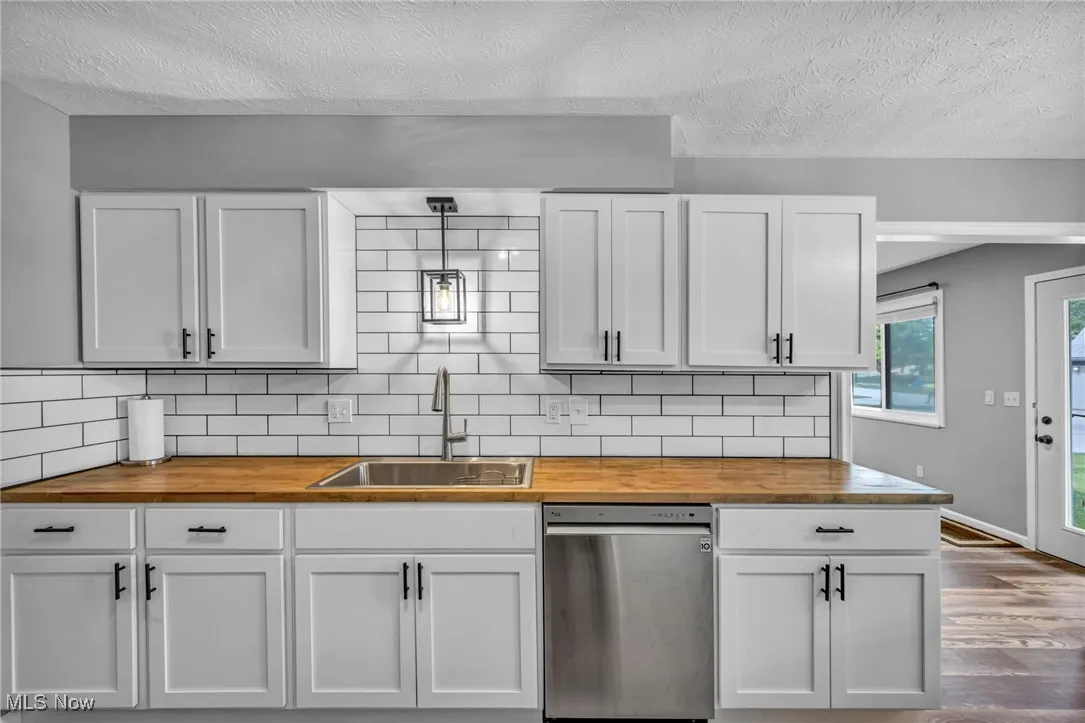 Kitchen with a textured ceiling, stainless steel dishwasher, tasteful backsplash, butcher block counters, and white cabinetry