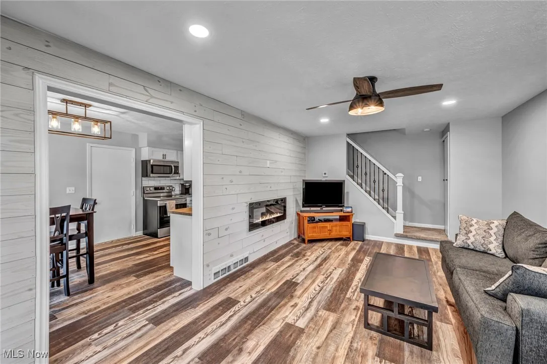 Living area featuring a glass covered fireplace, a ceiling fan, wood finished floors, wooden walls, and stairs