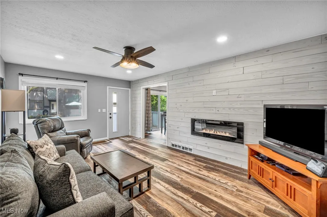 Living room featuring wooden walls, a textured ceiling, a glass covered fireplace, light wood-type flooring, and ceiling fan