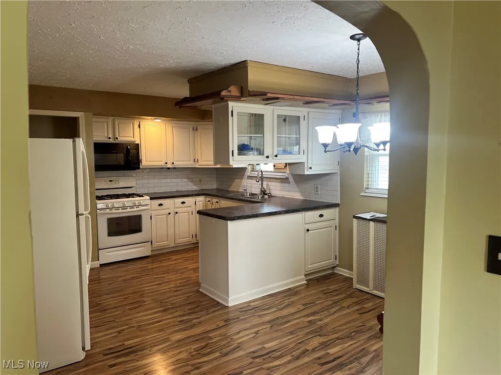 Kitchen featuring a peninsula, white appliances, dark countertops, dark wood-type flooring, and backsplash