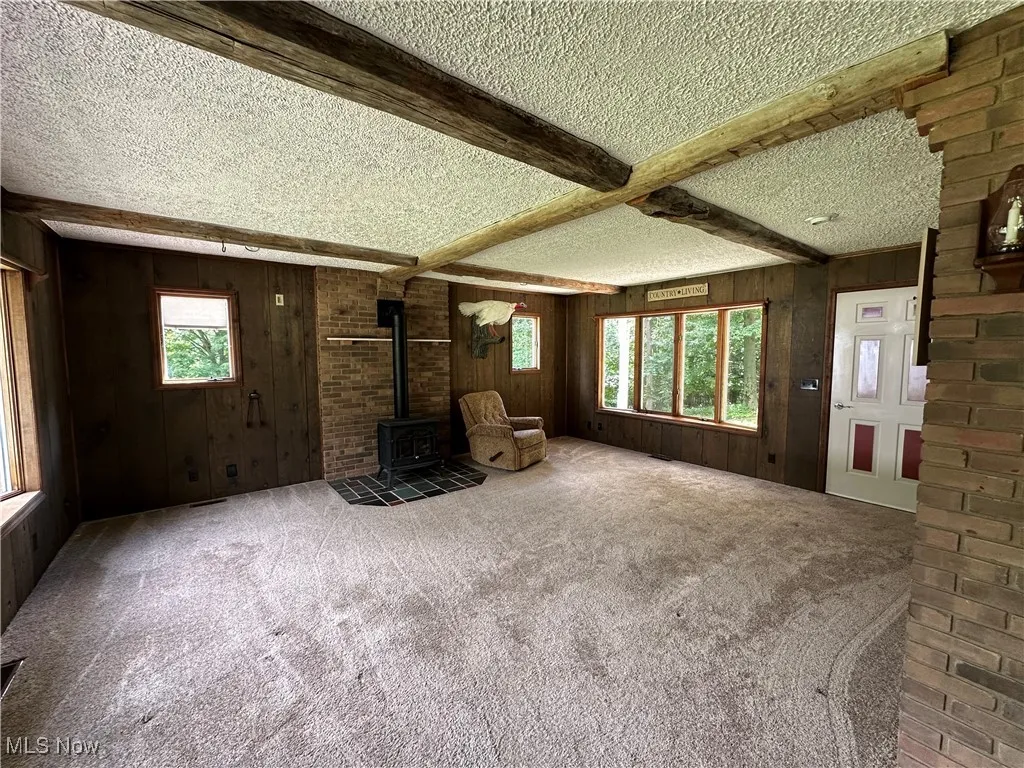  living room with beam ceiling, a wood stove, wooden walls, a textured ceiling, and carpet flooring