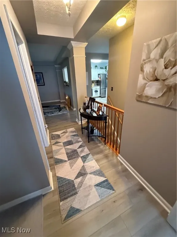Hallway featuring a textured ceiling, an upstairs landing, ornate columns, light wood-type flooring, and a tray ceiling
