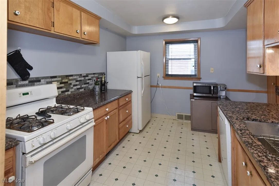 Kitchen with white appliances, a raised ceiling, dark stone countertops, and backsplash