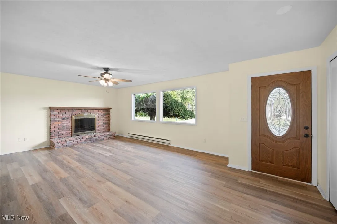 Unfurnished living room featuring a brick fireplace, a baseboard heating unit, light wood finished floors, and ceiling fan