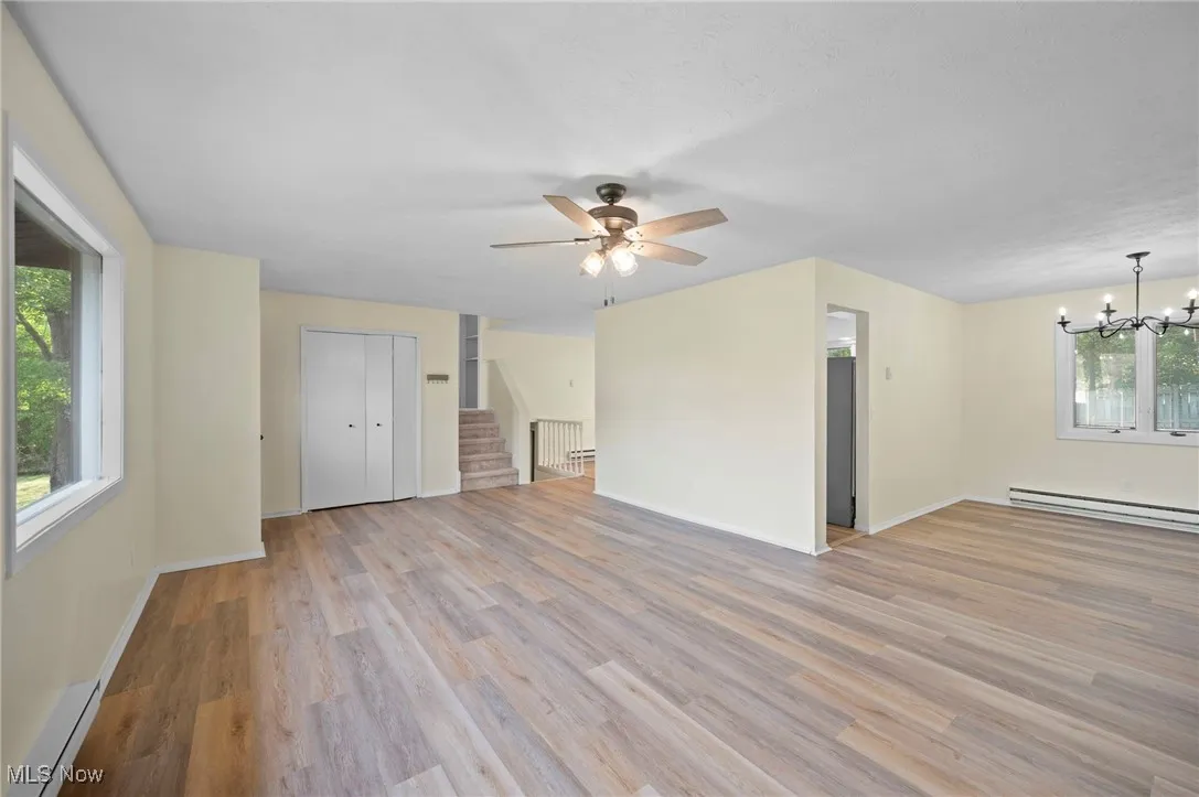 Unfurnished living room with stairs, a baseboard heating unit, light wood-style floors, a ceiling fan, and a chandelier