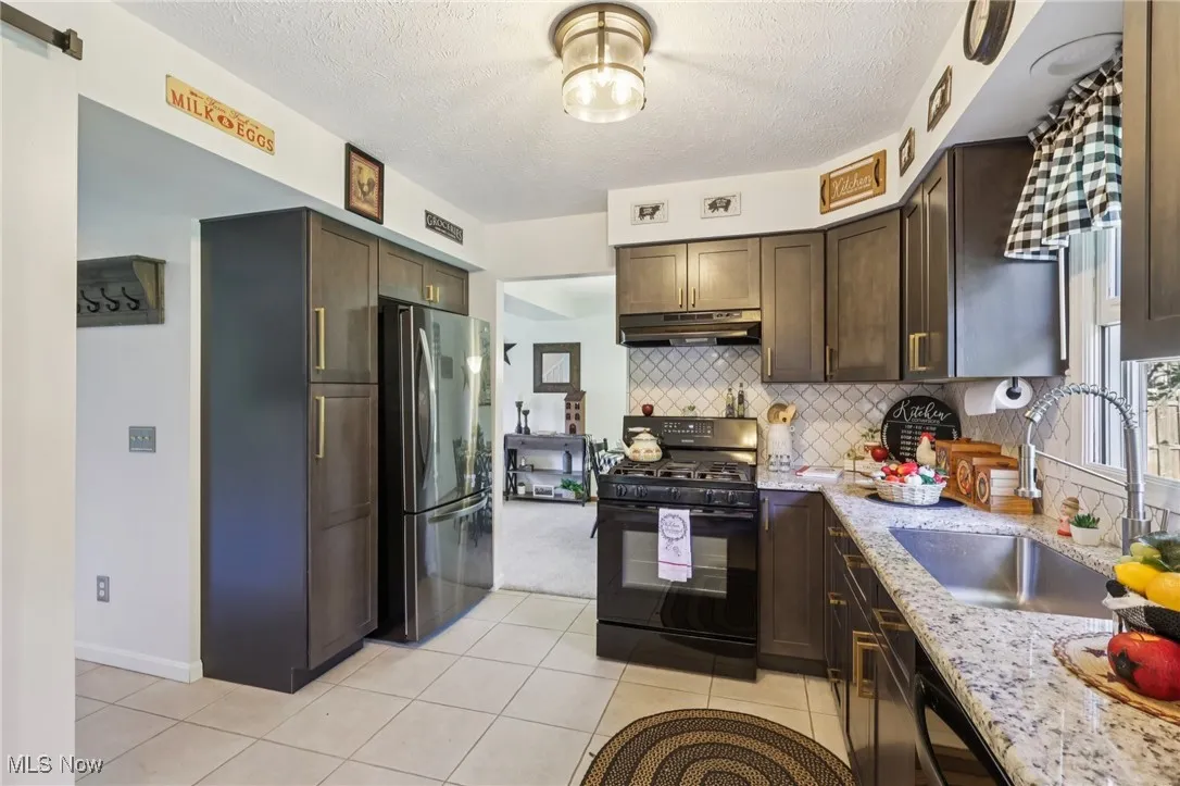 Kitchen with dark brown cabinets, black appliances, a textured ceiling, light stone counters, and decorative backsplash