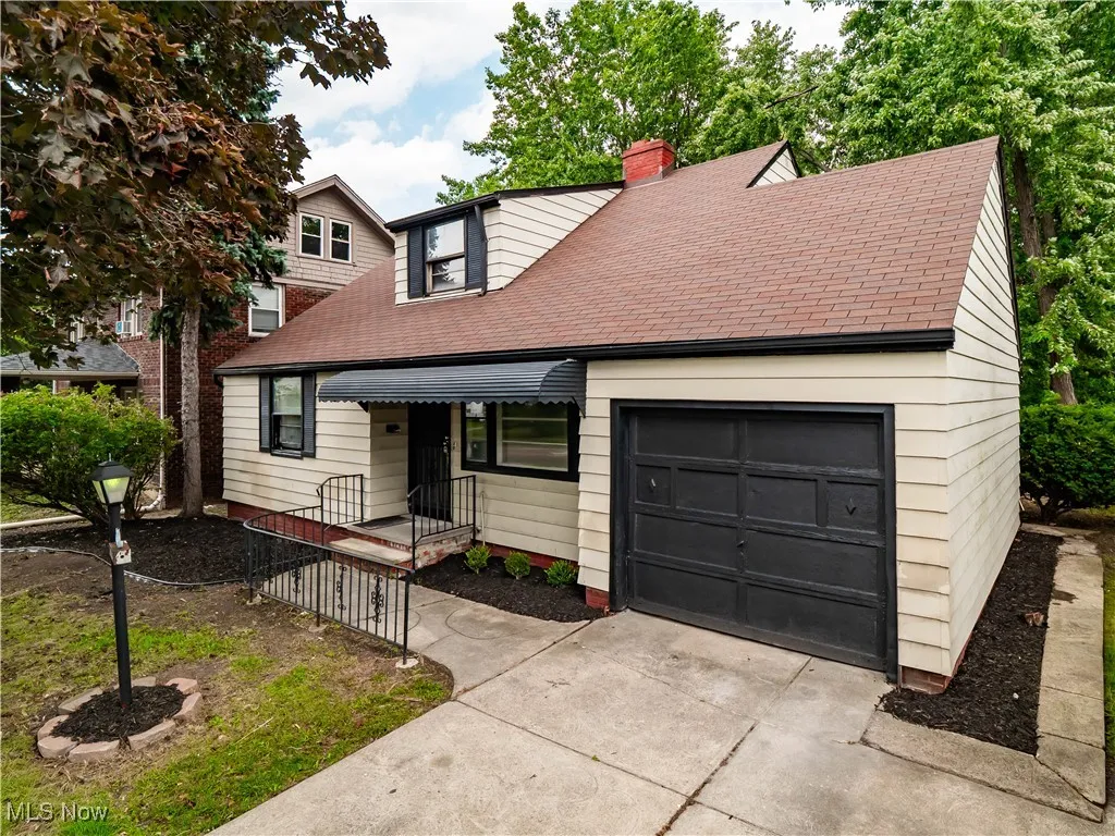 Cape cod home with a shingled roof, a garage, concrete driveway, and a chimney