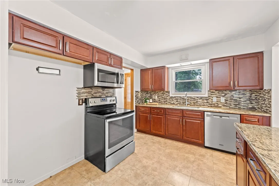 Kitchen featuring tasteful backsplash, appliances with stainless steel finishes, brown cabinetry, and light stone counters