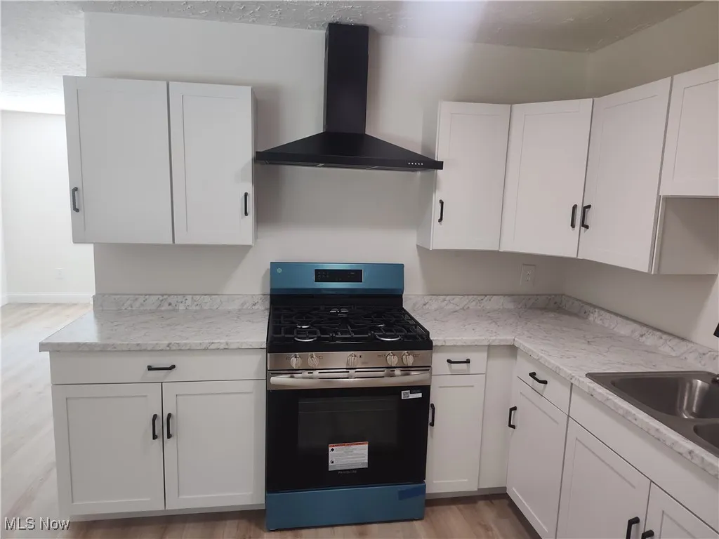 Kitchen featuring stainless steel range with gas cooktop, white cabinetry, wall chimney exhaust hood, light wood-style floors, and a textured ceiling