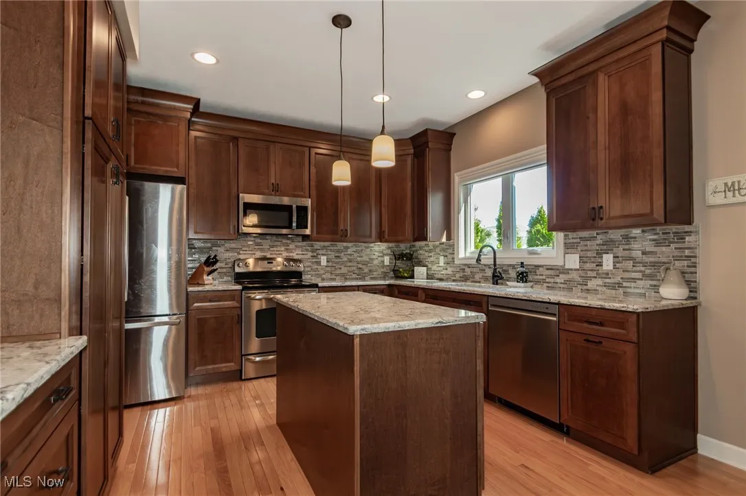 Kitchen featuring decorative light fixtures, stainless steel appliances, light wood-type flooring, a center island, and light stone countertops
