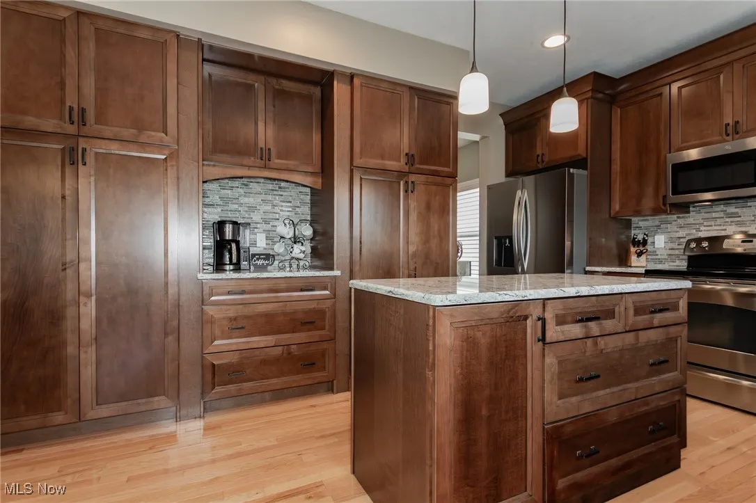 Kitchen featuring decorative backsplash, appliances with stainless steel finishes, pendant lighting, light stone counters, and light wood-style flooring