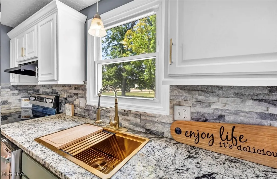 Kitchen with backsplash, light stone counters, white cabinets, decorative light fixtures, and stainless steel dishwasher