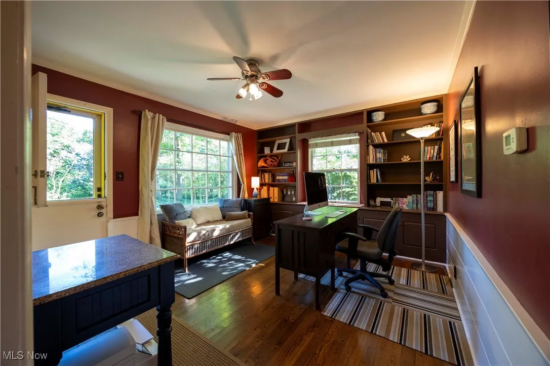 Office space featuring dark wood-style floors, ornamental molding, a ceiling fan, built in shelves, and a wainscoted wall