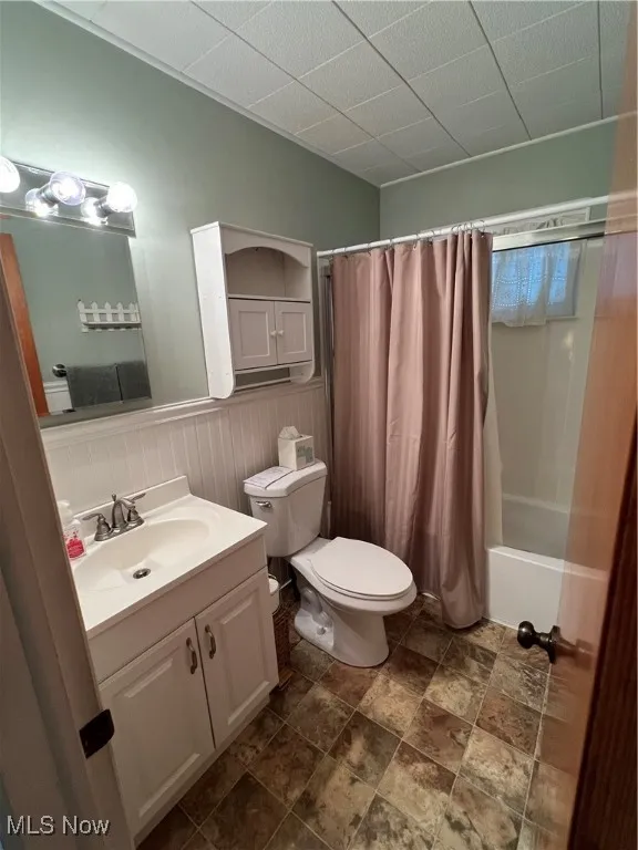 Bathroom featuring shower / bath combination with curtain, vanity, a paneled ceiling, a wainscoted wall, and dark stone finish​​‌​​​​‌​​‌‌​‌‌​​​‌‌​‌​‌​‌​​​‌​​ flooring