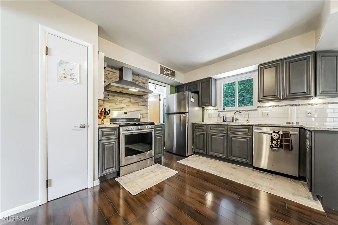 Kitchen featuring appliances with stainless steel finishes, wall chimney range hood, backsplash, light stone countertops, and dark wood-type flooring