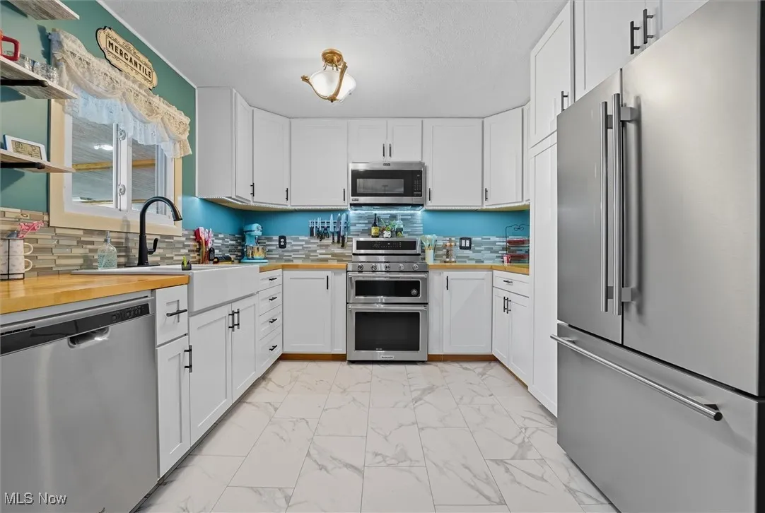 Kitchen with appliances with stainless steel finishes, white cabinets, light marble finish flooring, backsplash, and butcher block countertops