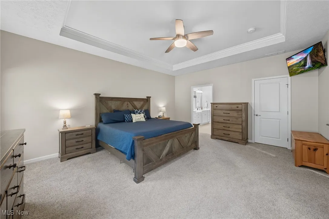 Bedroom with a raised ceiling, light colored carpet, ceiling fan, and ensuite bathroom