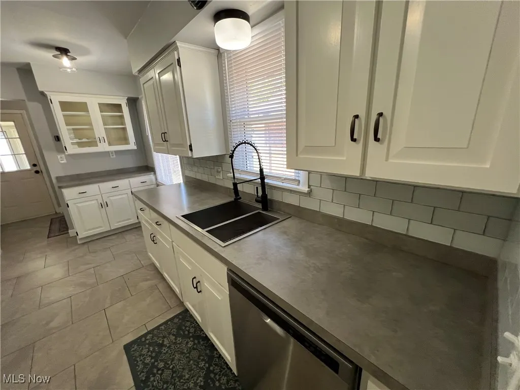Kitchen featuring white cabinetry, stainless steel dishwasher, decorative backsplash, light tile patterned floors, and glass insert cabinets