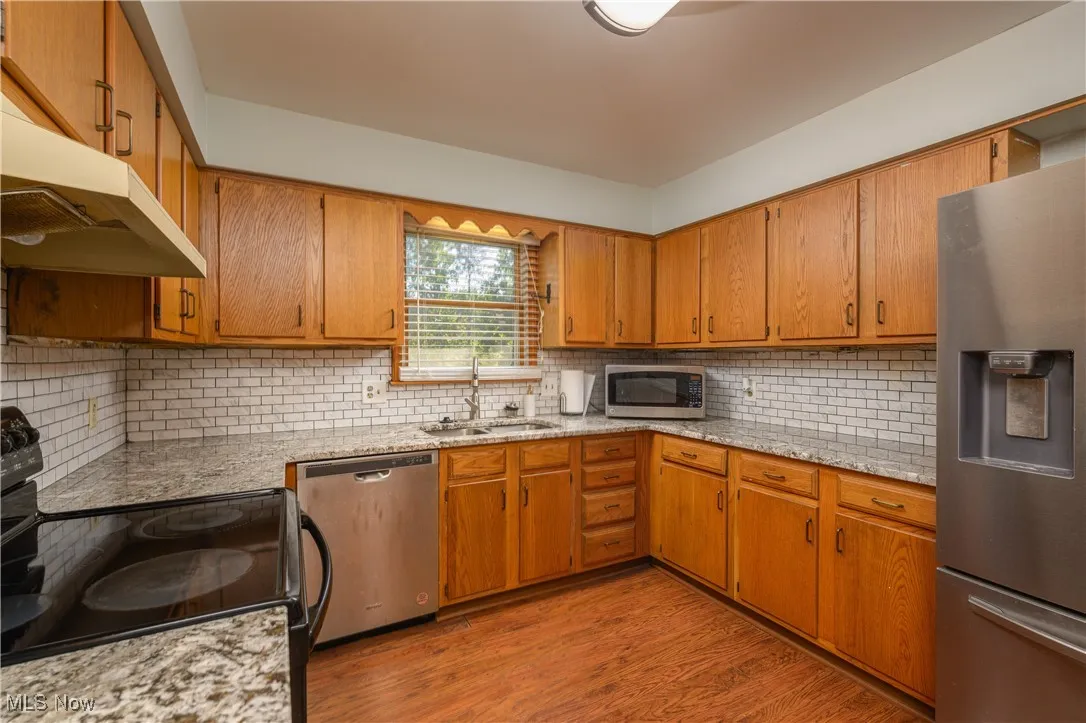 Kitchen with stainless steel appliances, tasteful backsplash, light wood finished floors, light stone countertops, and brown cabinetry
