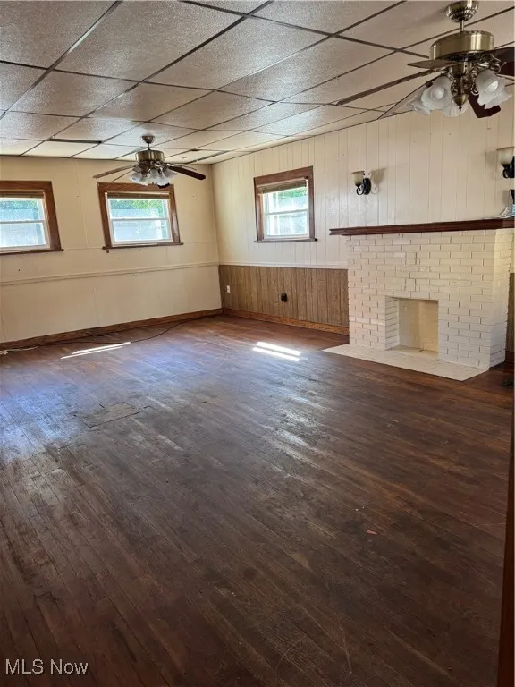 Unfurnished living room featuring a ceiling fan, wood walls, dark wood-style flooring, a brick fireplace, and a drop ceiling