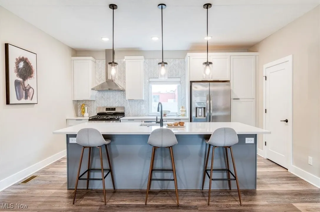 Kitchen featuring wall chimney range hood, appliances with stainless steel finishes, a kitchen island with sink, white cabinets, and recessed lighting