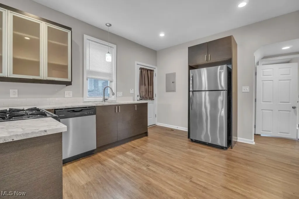 Kitchen featuring appliances with stainless steel finishes, light wood finished floors, light stone counters, hanging light fixtures, and dark brown cabinetry