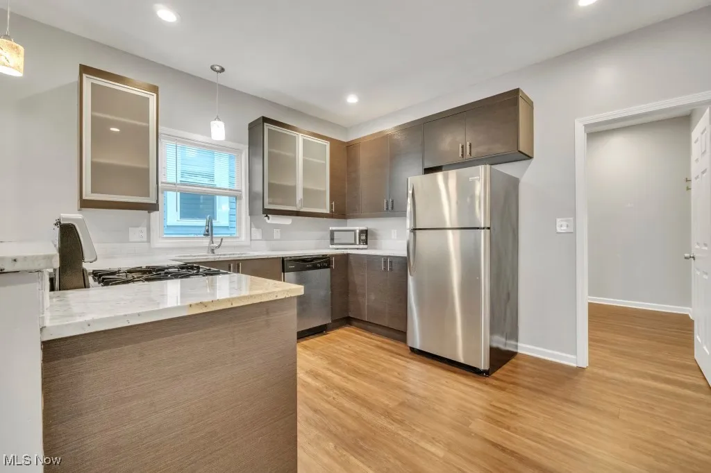 Kitchen with glass insert cabinets, stainless steel appliances, light wood-type flooring, light stone counters, and hanging light fixtures