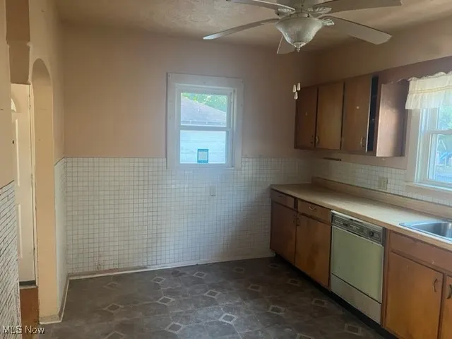 Kitchen with wainscoting, tile walls, light countertops, stainless steel dishwasher, and ceiling fan