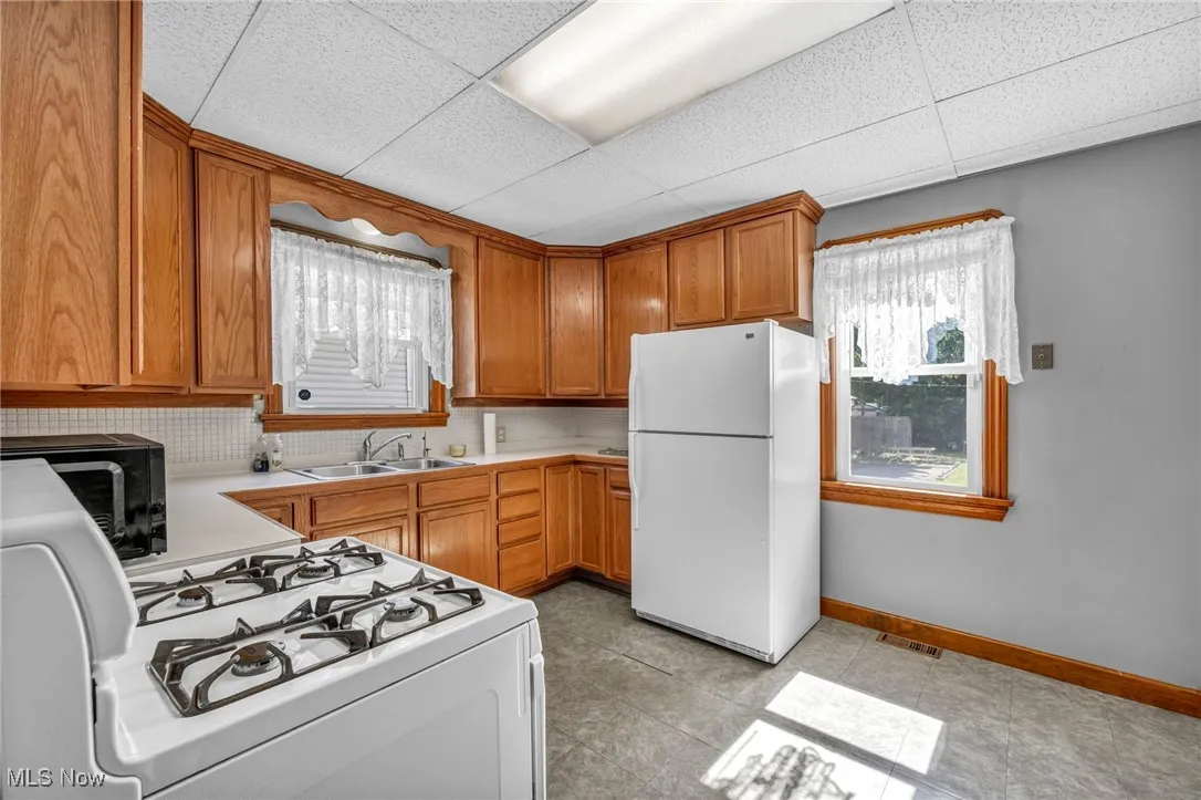 Kitchen featuring white appliances, brown cabinetry, decorative backsplash, a drop ceiling, and light tile patterned flooring