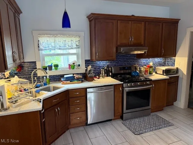 Kitchen with appliances with stainless steel finishes, backsplash, dark brown cabinets, under cabinet range hood, and hanging light fixtures