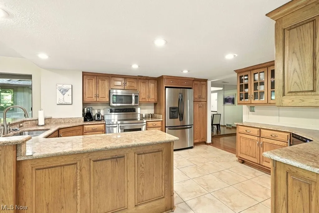 Kitchen with appliances with stainless steel finishes, brown cabinets, glass insert cabinets, light tile patterned flooring, and recessed lighting