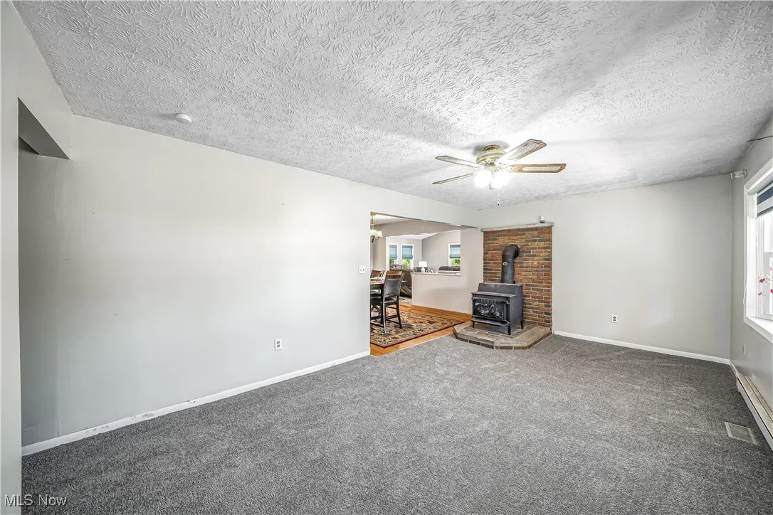 Unfurnished living room featuring a wood stove, carpet, a ceiling fan, and a textured ceiling