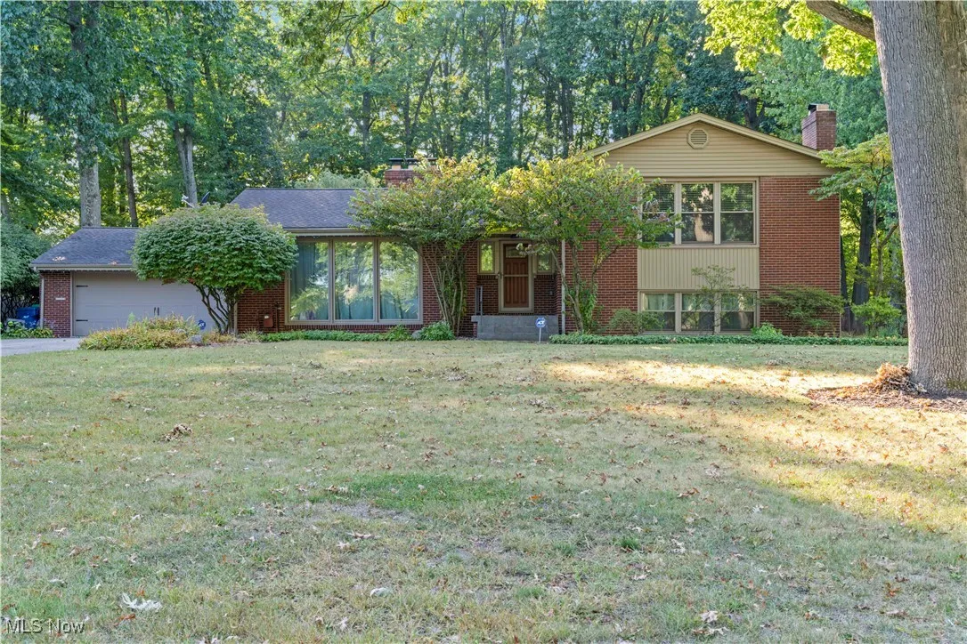 Split level home featuring a chimney, a front lawn, brick siding, an attached garage, and view of wooded area