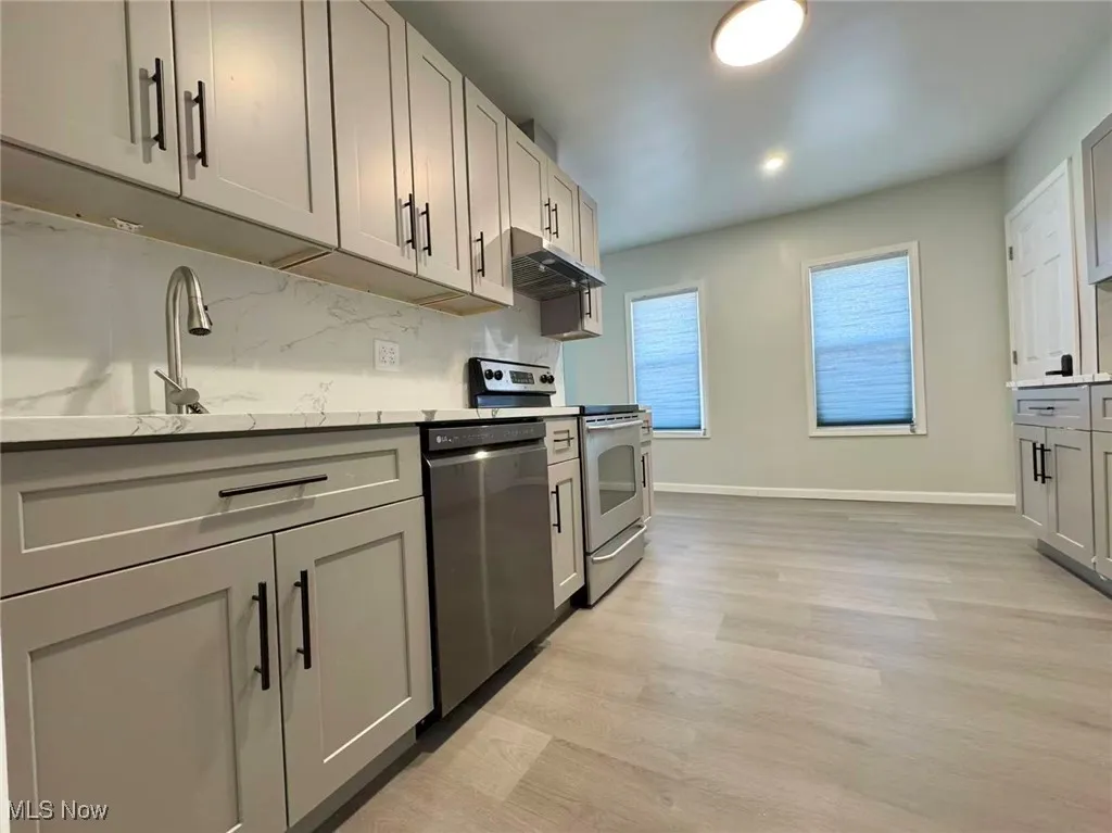 Kitchen with gray cabinetry, appliances with stainless steel finishes, under cabinet range hood, light wood-style floors, and light stone countertops