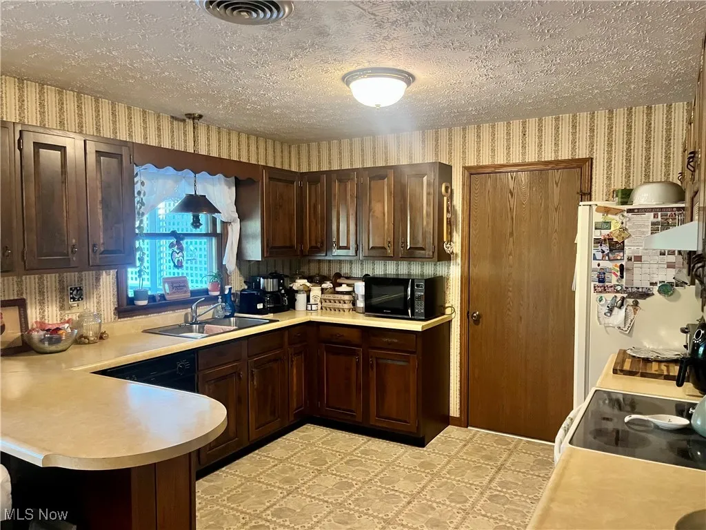 Kitchen featuring wallpapered walls, dark brown cabinets, light countertops, white appliances, and a textured ceiling