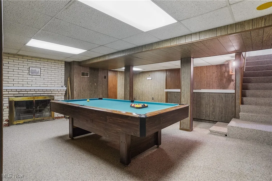 Playroom with carpet flooring, a brick fireplace, wooden walls, billiards, and a drop ceiling