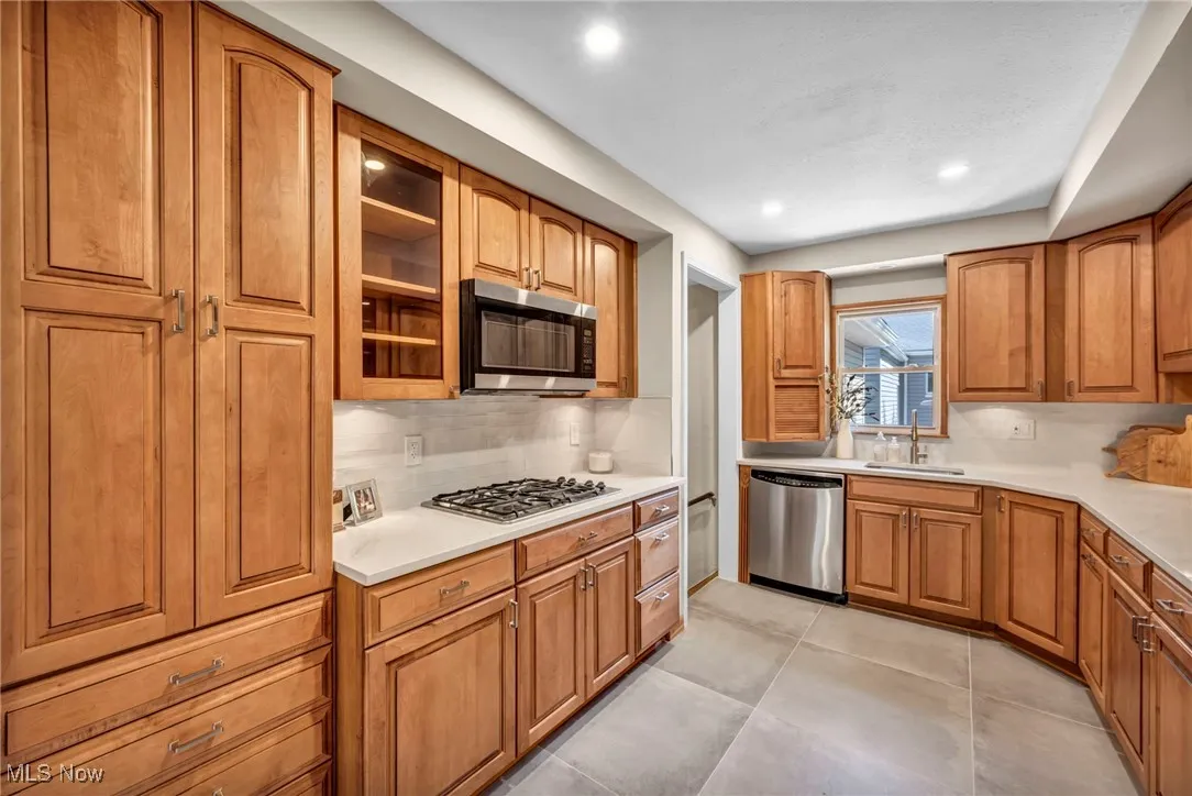 Kitchen with brown cabinets, appliances with stainless steel finishes, tasteful backsplash, glass insert cabinets, and light tile patterned floors