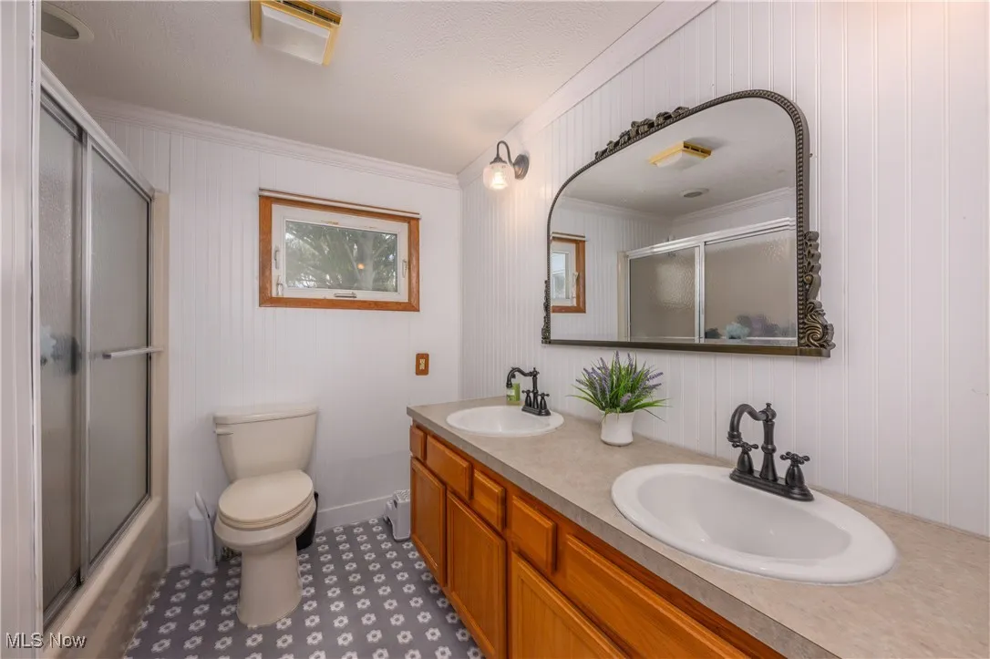 Bathroom featuring double vanity, crown molding, bath / shower combo with glass door, and wood walls