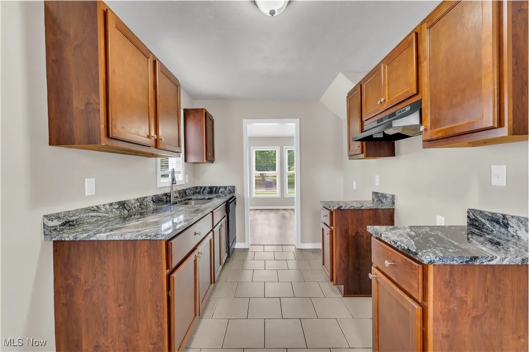Kitchen featuring brown cabinets, dark stone counters, under cabinet range hood, and light tile patterned flooring
