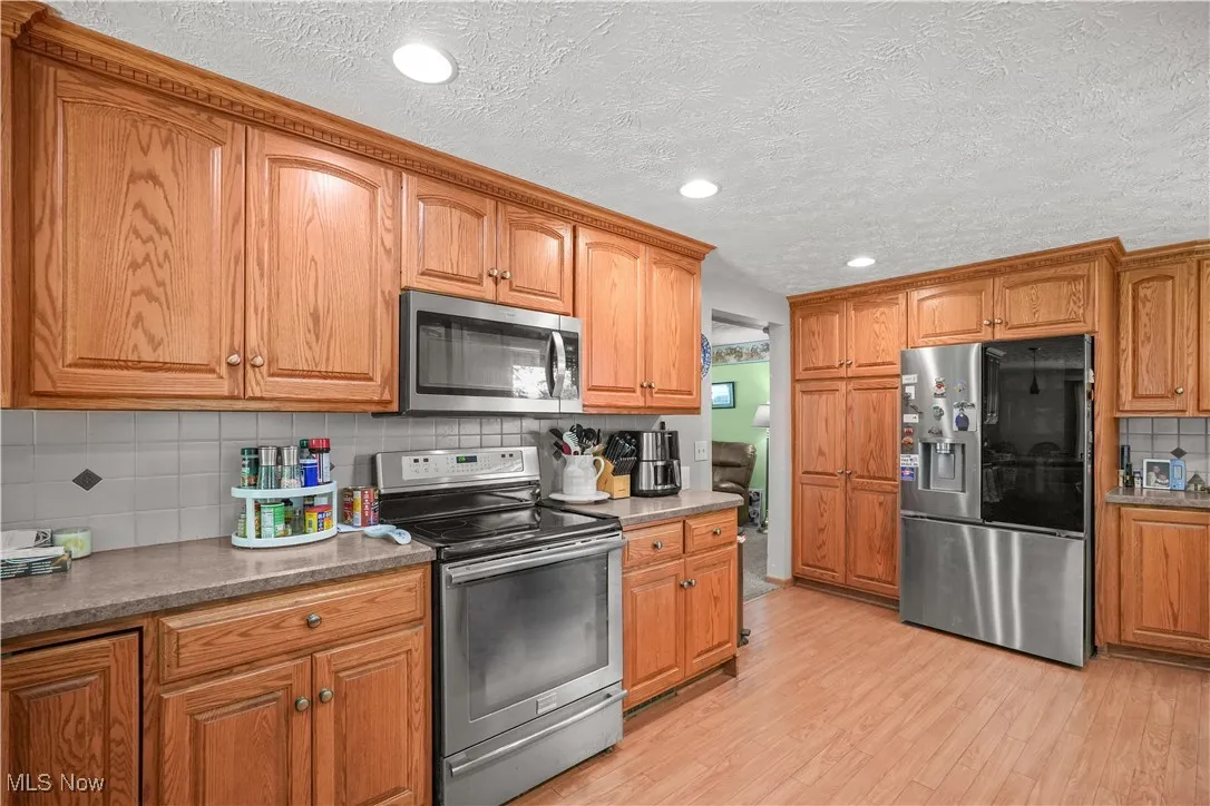 Kitchen featuring backsplash, stainless steel appliances, a textured ceiling, brown cabinets, and light wood-type flooring