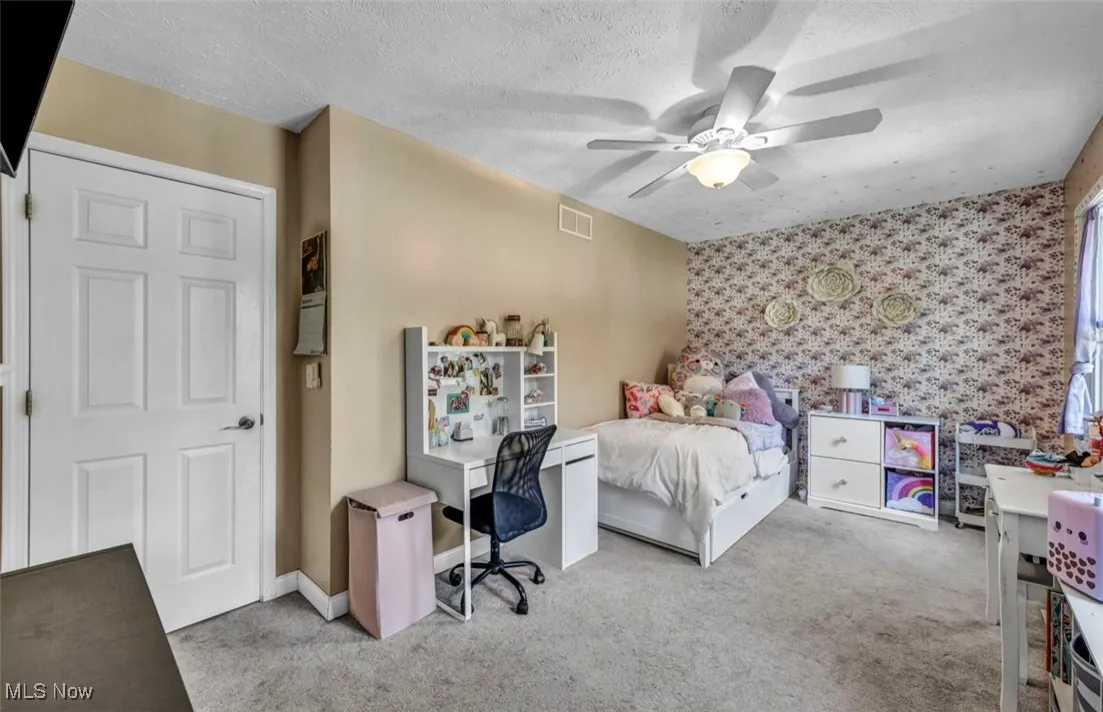 Bedroom featuring an accent wall, light carpet, a textured ceiling, wallpapered walls, and ceiling fan