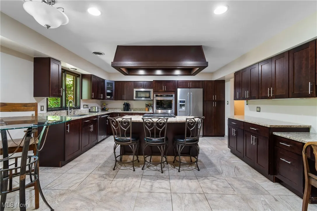 Kitchen with a kitchen breakfast bar, wall chimney range hood, a center island, stainless steel appliances, and dark brown cabinets