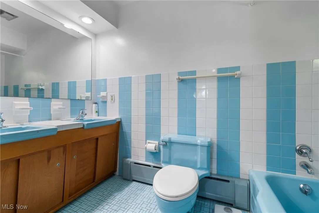 Full bathroom with tile walls, a baseboard radiator, double vanity, a wainscoted wall, and light tile patterned flooring