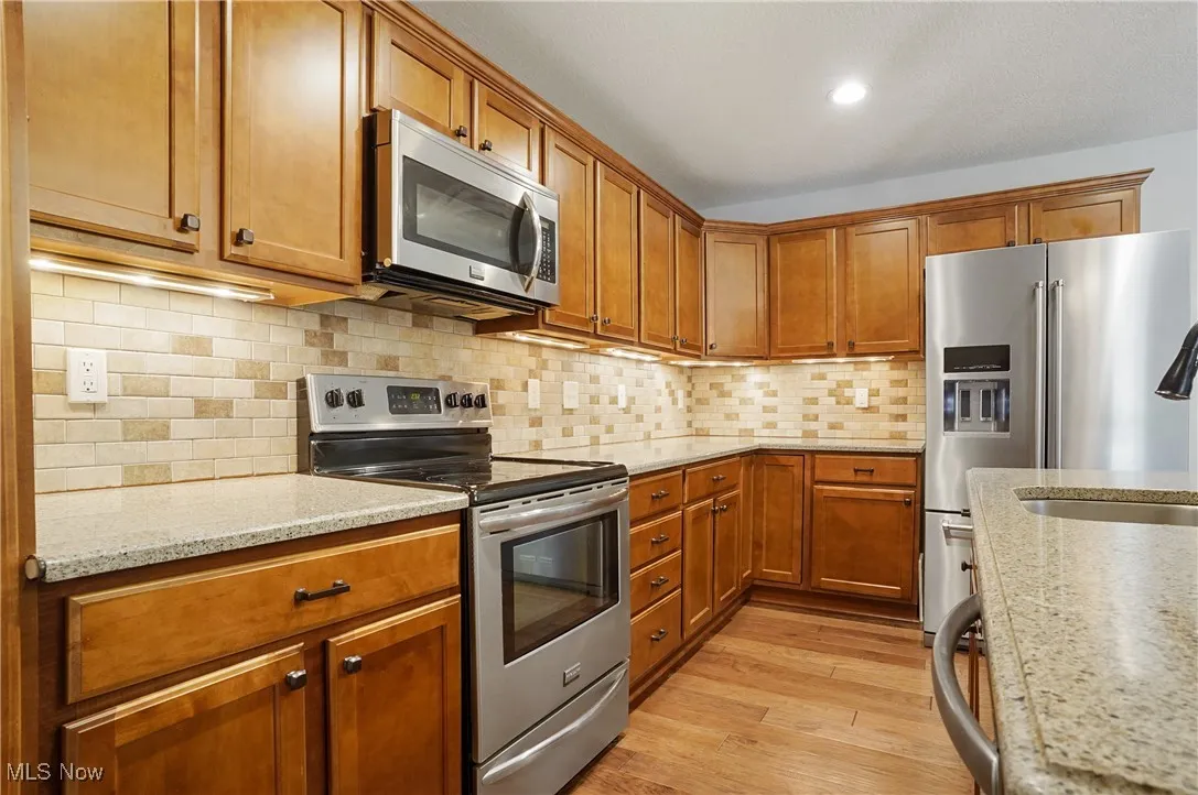 Kitchen with appliances with stainless steel finishes, brown cabinetry, decorative backsplash, light stone countertops, and light wood finished floors