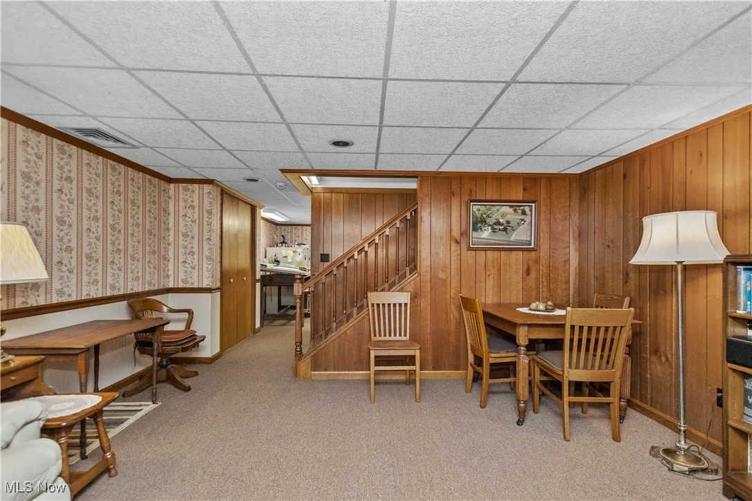 Dining room featuring light carpet, stairway, wooden walls, and a paneled ceiling