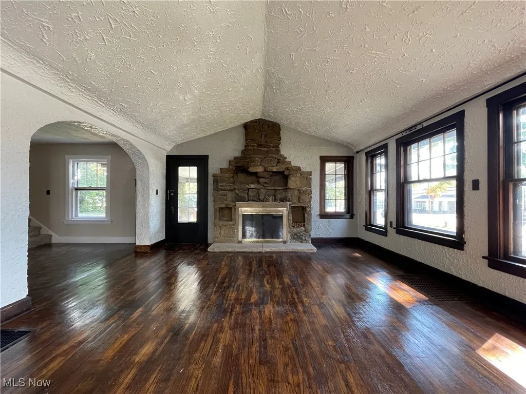 Unfurnished living room featuring a textured wall, dark wood finished floors, a textured ceiling, vaulted ceiling, and arched walkways