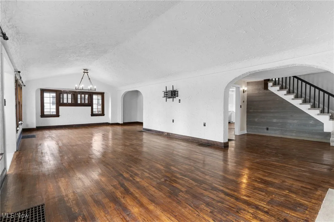 Unfurnished living room with dark wood finished floors, a chandelier, lofted ceiling, a textured wall, and arched walkways