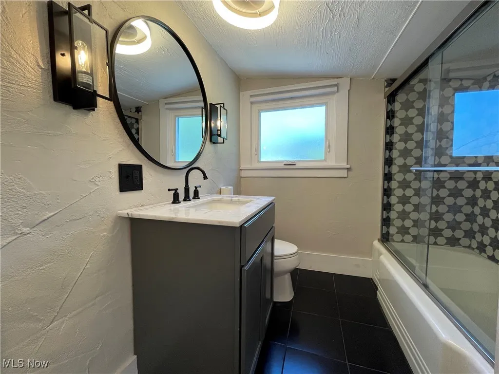 Bathroom with bath / shower combo with glass door, dark tile patterned flooring, vanity, a textured ceiling, and a textured wall
