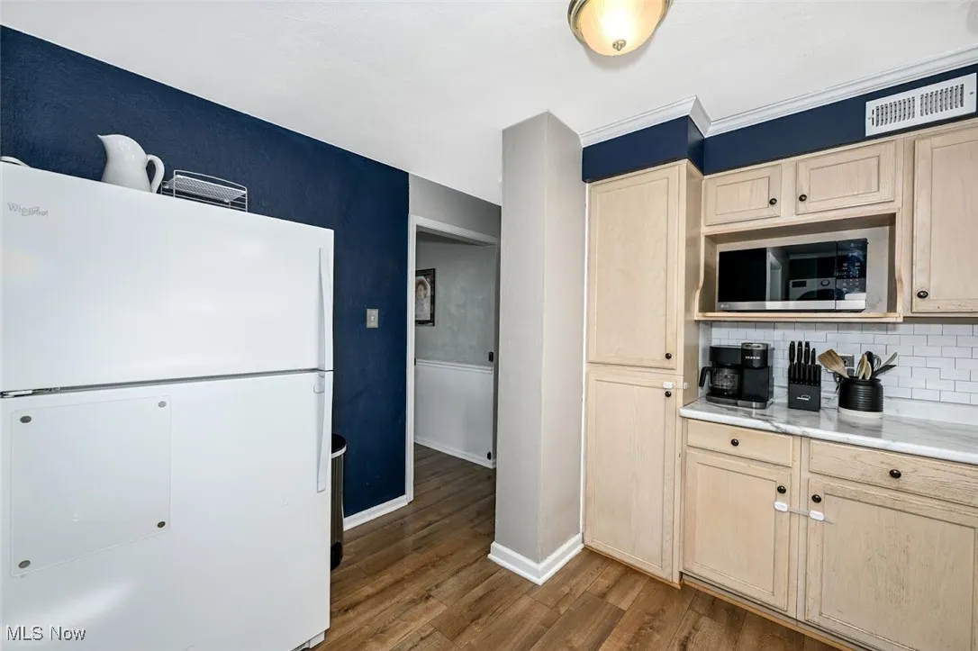 Kitchen with white fridge, light brown cabinetry, dark wood-type flooring, backsplash, and stainless steel microwave